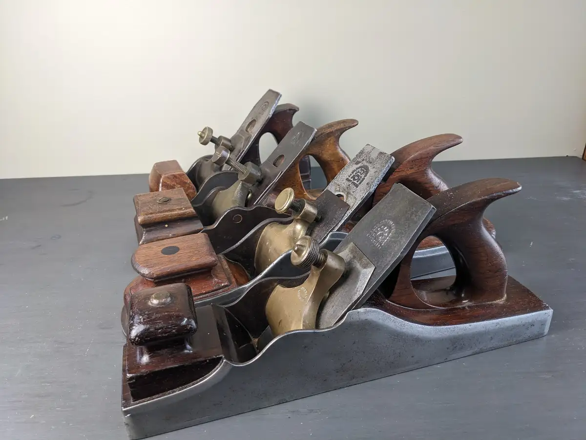 A row of vintage hand planes with wooden handles and metal blades are neatly arranged in a metal stand on a dark gray…