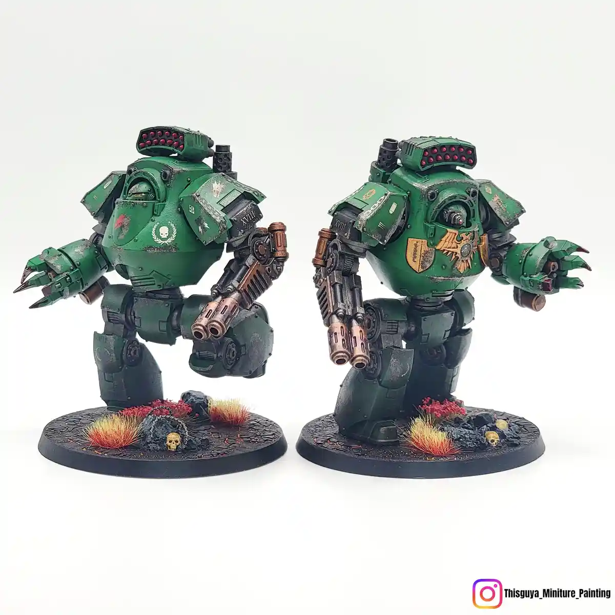 Two identical green, heavily armored, bipedal war machines stand side-by-side on circular bases with painted terrain. The…