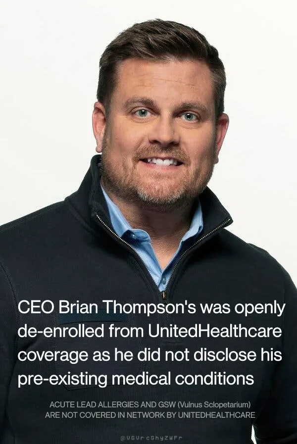 A man with short brown hair and a beard, wearing a black zip-up jacket over a blue collared shirt, smiles at the camera…