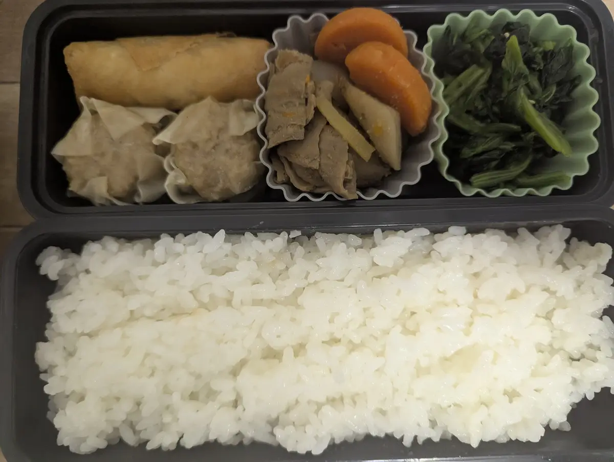 a top-down photo of a black bento box filled with white rice in the bottom compartment and three smaller compartments above…