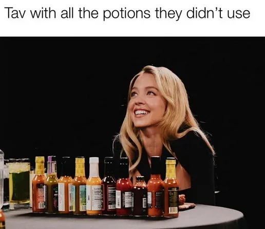 A blonde woman smiles upward while seated at a table with a row of various hot sauce bottles arranged in front of her. The…