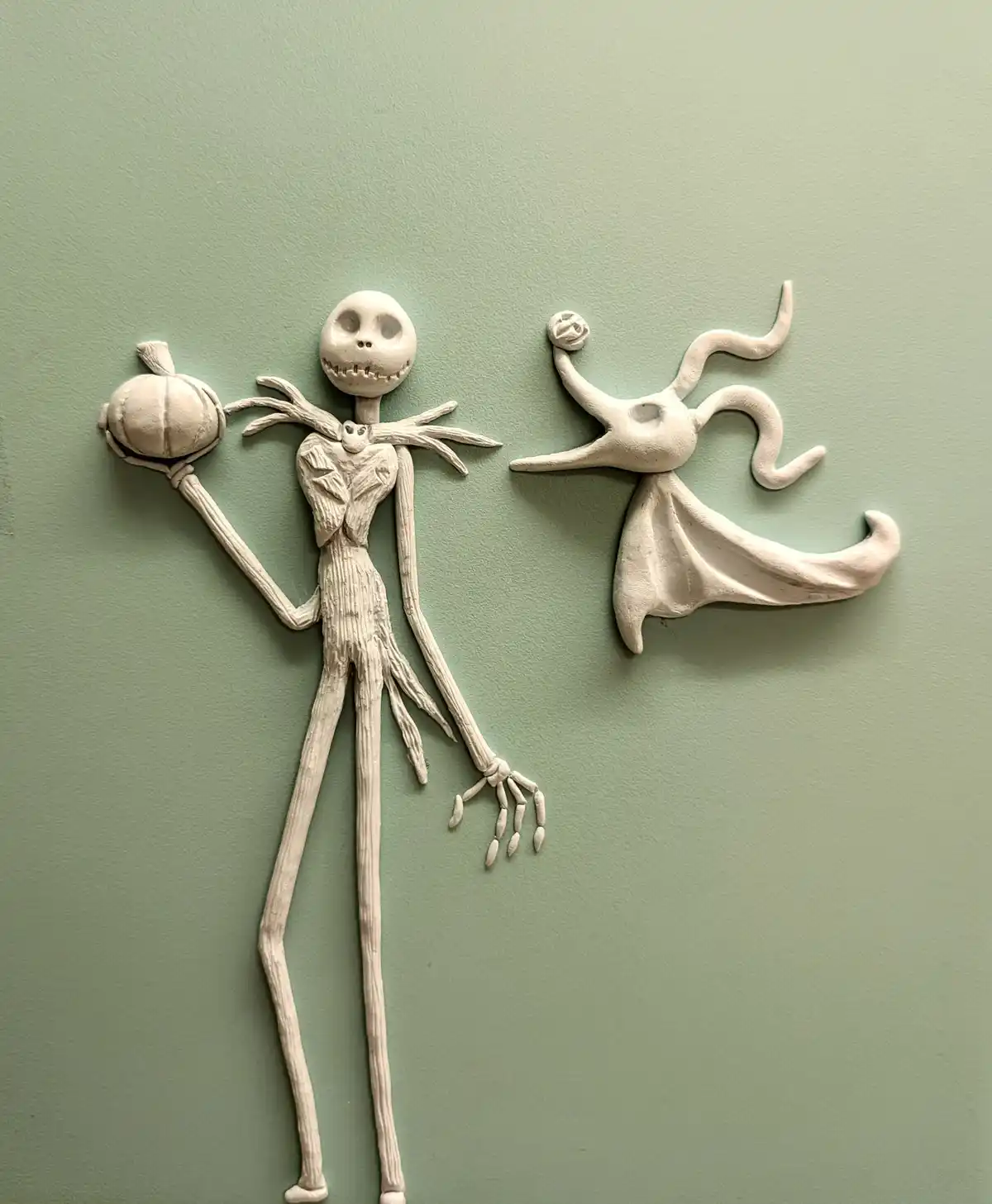 Two white clay sculptures of Jack Skellington and Zero are mounted on a pale green wall. Jack holds a pumpkin in one hand…