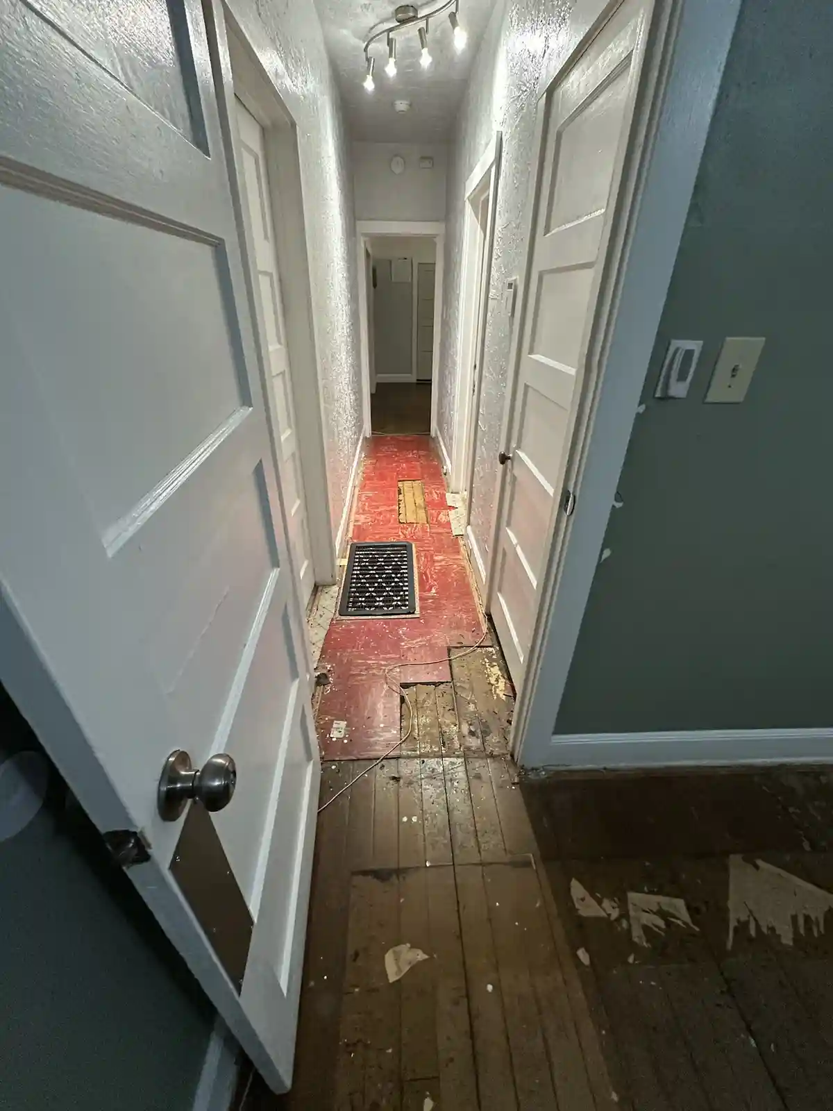 A narrow hallway in a residential apartment shows a chaotic floor with partially removed red flooring, exposed wooden…
