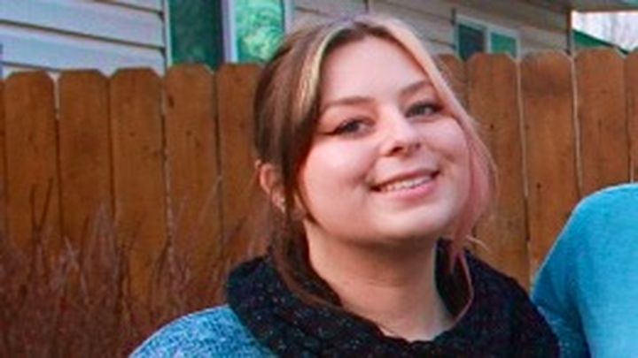 A young woman with light brown hair and a slight smile stands outdoors in front of a wooden fence, wearing a blue sweater…