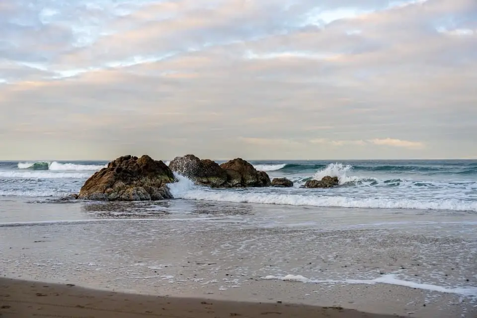 A serene beach scene with waves crashing against large rocks, under a cloudy sky with soft light. Overlaid text reads: 'i'm…