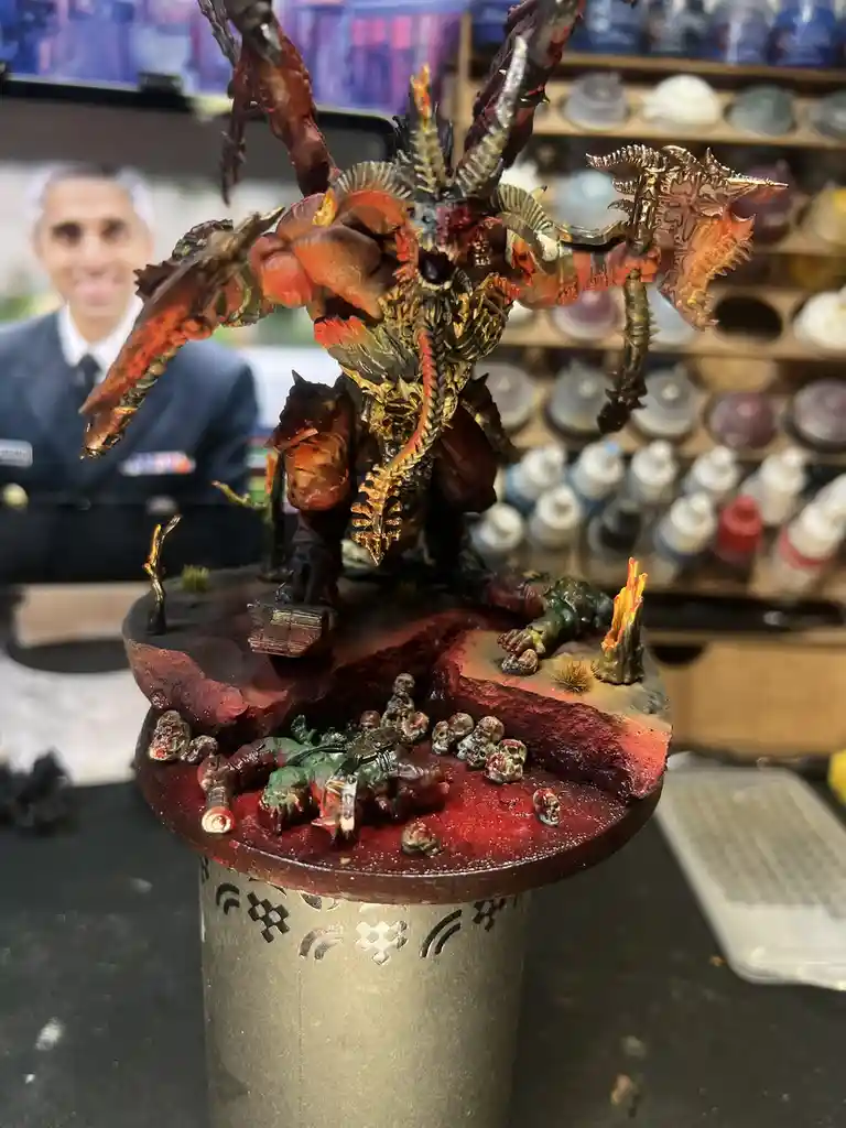 A highly detailed, painted fantasy demon miniature with multiple arms, horns, and weapons stands on a circular base littered…