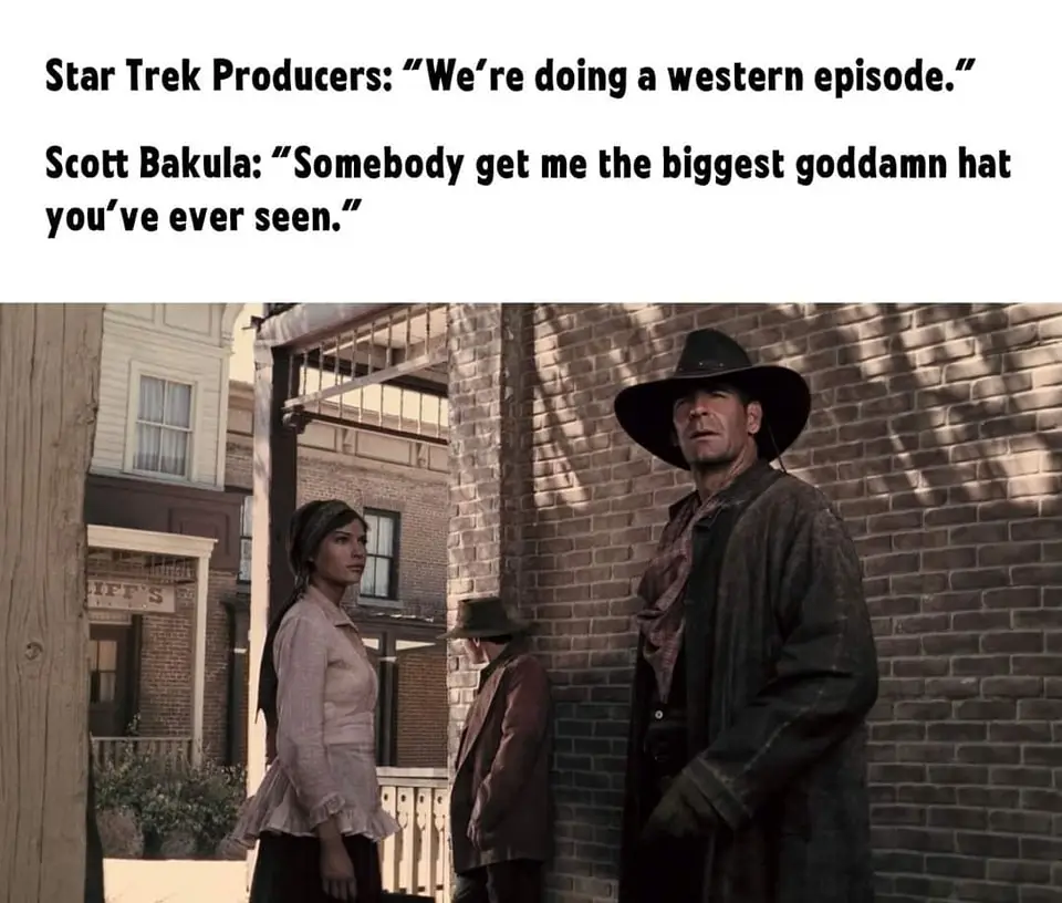 The image shows a scene from a Western movie or TV show with three characters in a dusty town street. A man in a large…