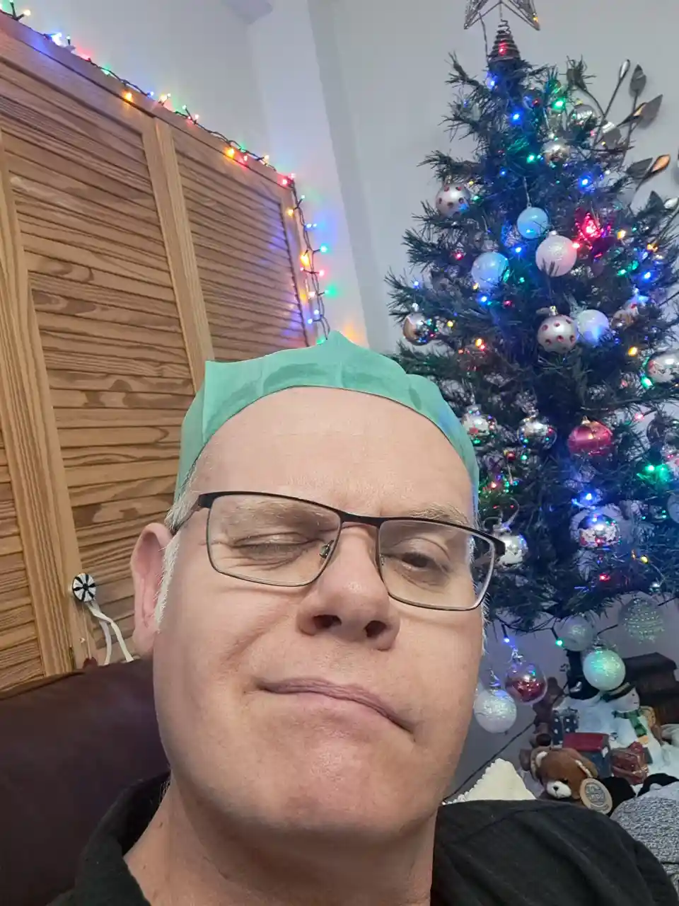 A man with glasses and a green surgical cap sits in front of a decorated Christmas tree with colorful lights and ornaments…