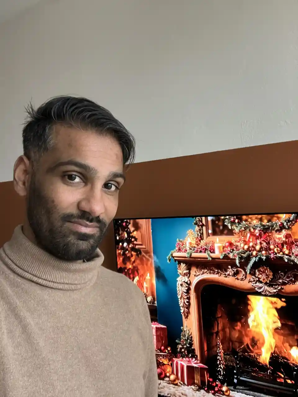 A man in a beige turtleneck sweater stands in front of a TV displaying a festive fireplace scene with ornaments, candles…