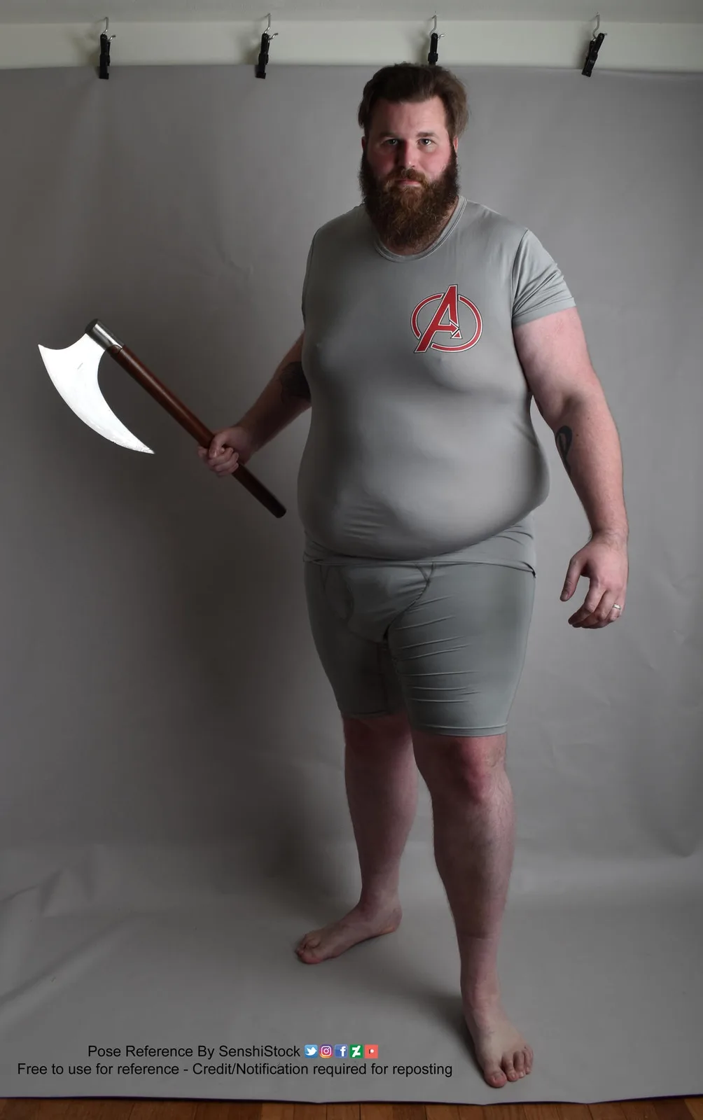 A man with a full beard and tattoos stands barefoot against a gray backdrop, wearing a tight gray t-shirt with a red…