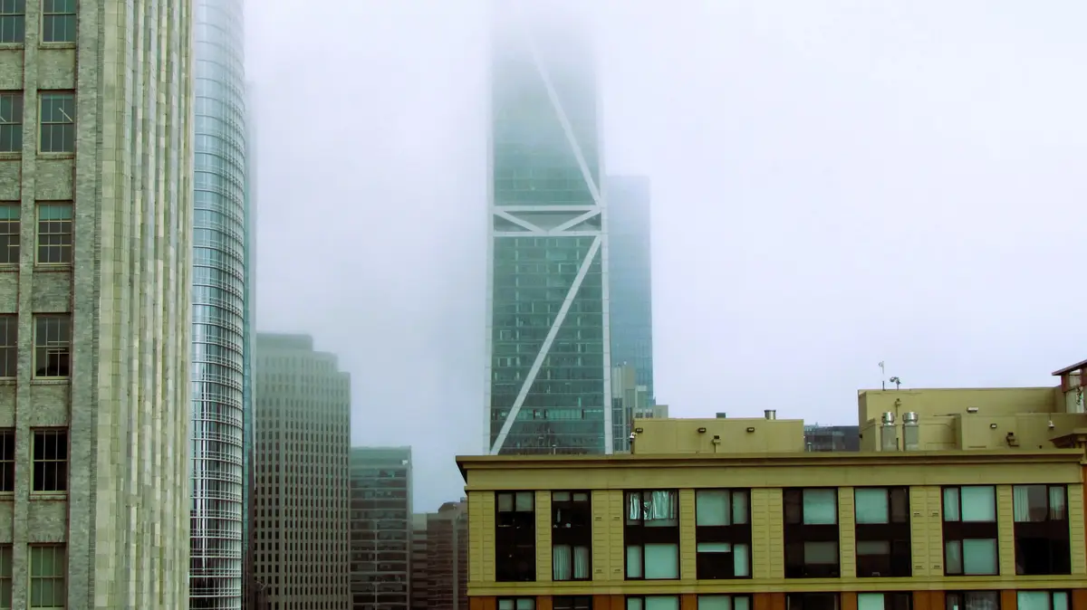 A cityscape photo shows a tall glass skyscraper partially obscured by thick fog, surrounded by other buildings. The overlaid…