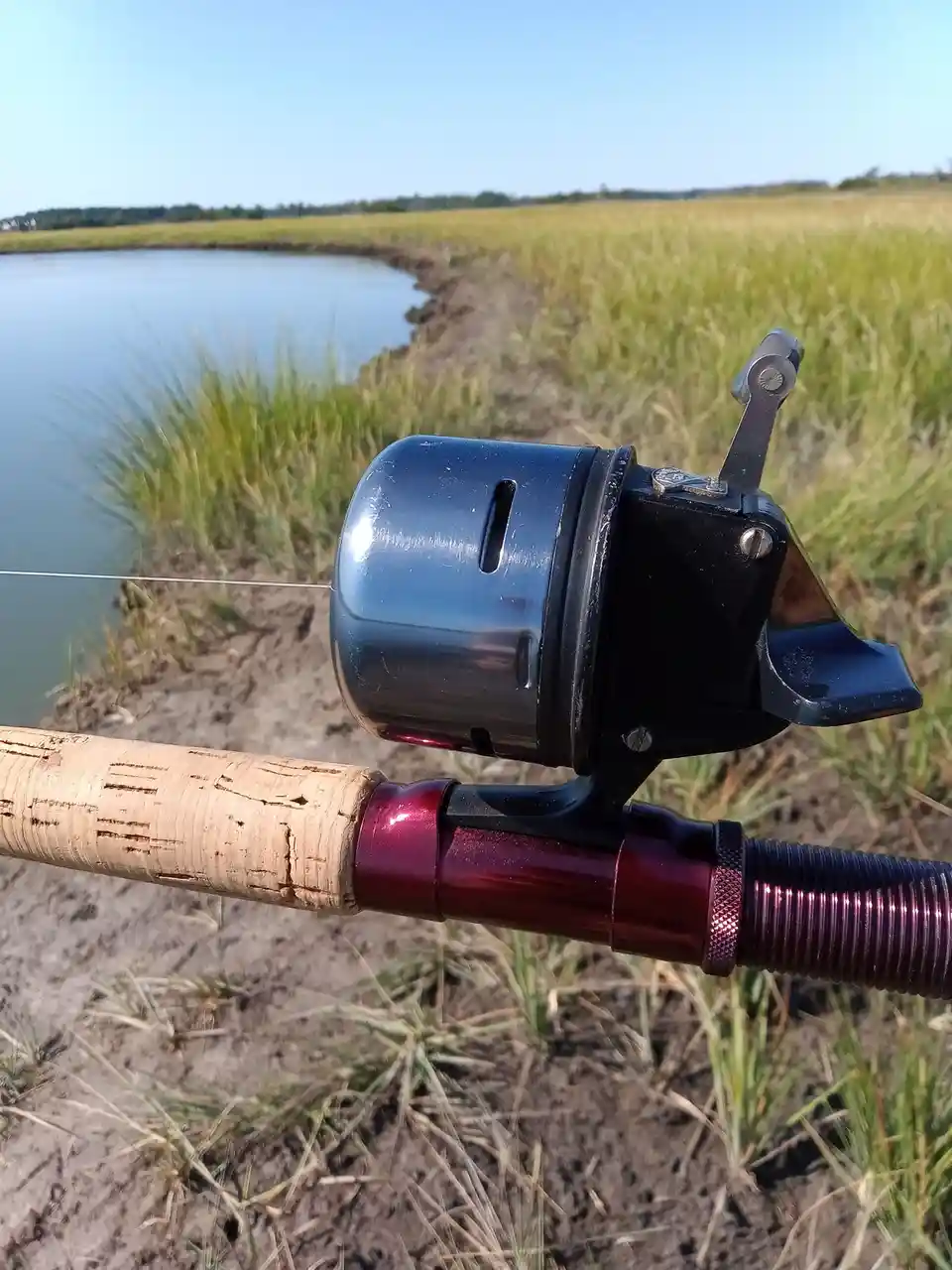 A close-up photo of a fishing rod with a metallic reel and cork handle, set against a backdrop of a calm body of water and…