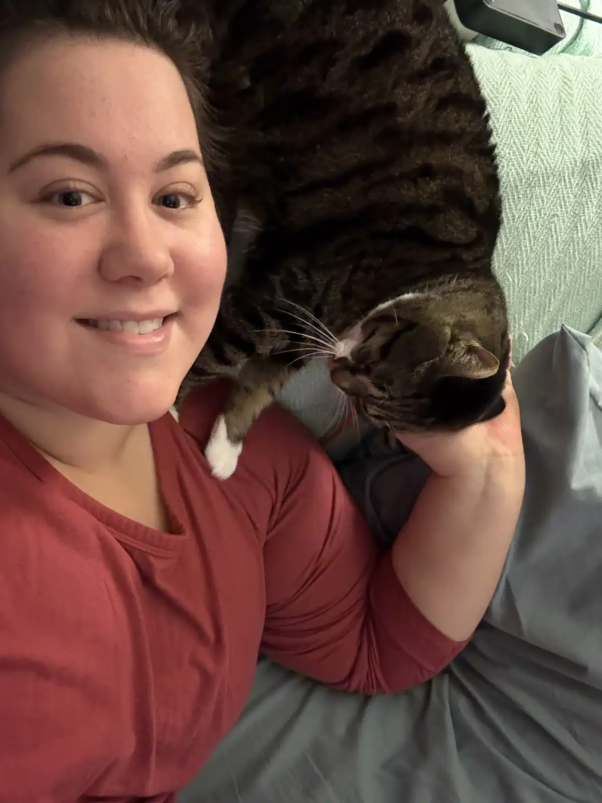 A person in a red shirt smiles at the camera while holding a large, dark tabby cat that is curled up and resting on their…
