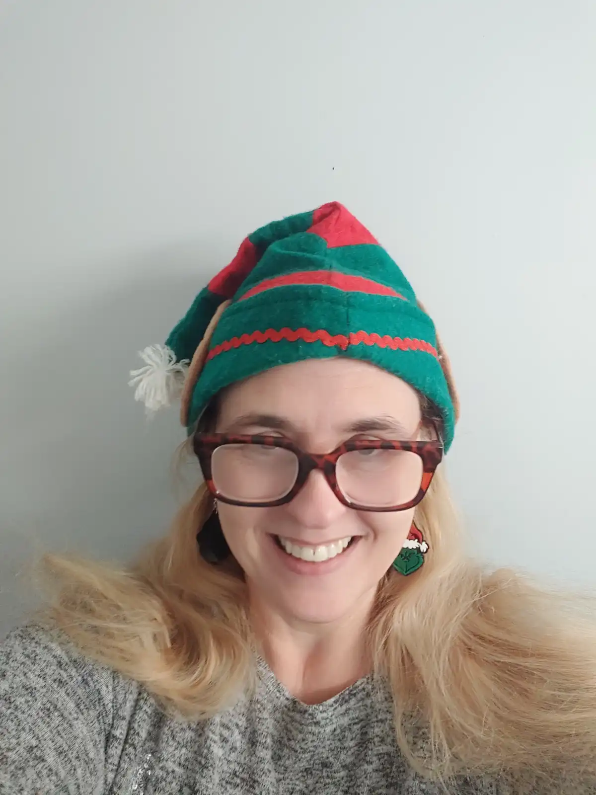 A person with blonde hair and glasses wears a green and red striped elf hat with a white pom-pom and festive earrings…