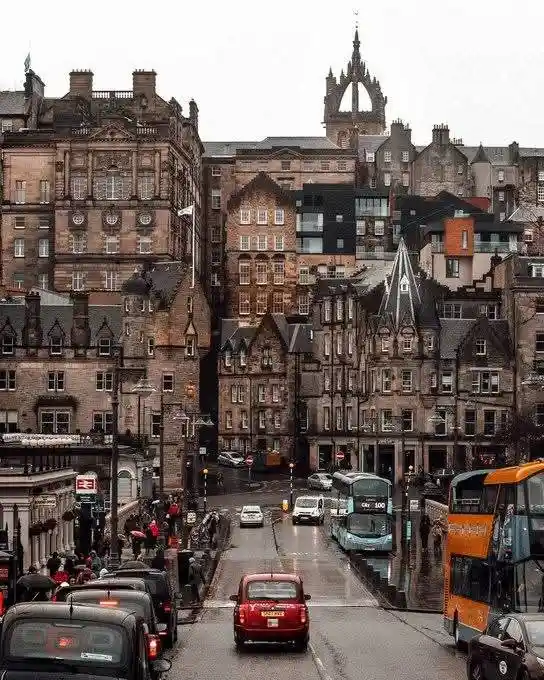 A street view of a historic city with old stone buildings and a prominent spire in the background. A modern, dark-colored…