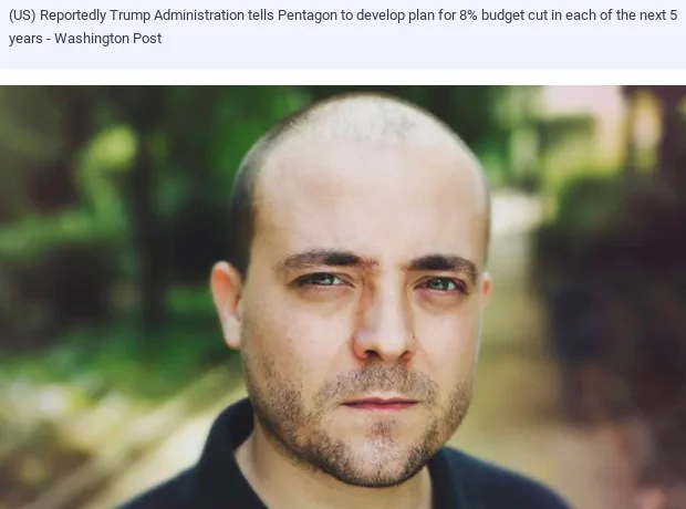 A close-up photo of a man with a shaved head and light stubble, looking directly at the camera with a serious, slightly…