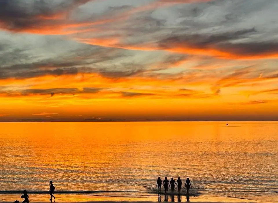 A serene beach sunset with vibrant orange and red clouds reflected in calm water, silhouetting several people walking along…