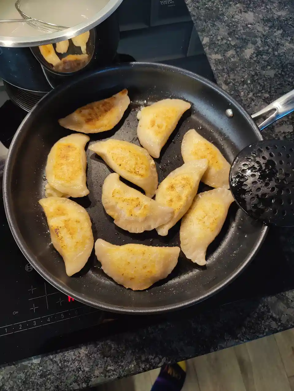 A black frying pan on a stovetop contains several golden-brown, crescent-shaped dumplings being fried. A black slotted…
