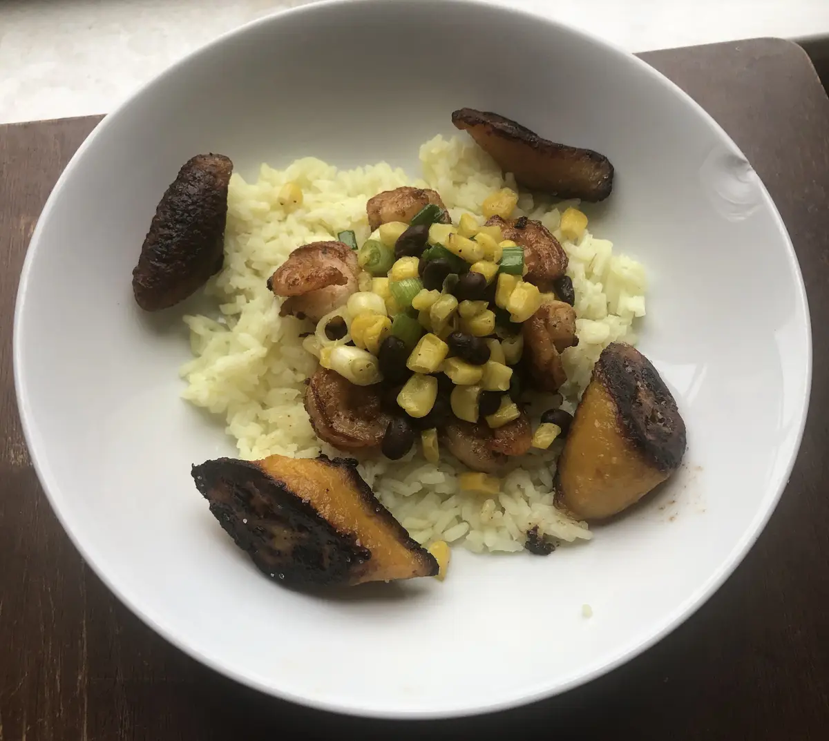 A white bowl contains a meal of rice topped with shrimp, corn, black beans, and chopped green onions, surrounded by several…