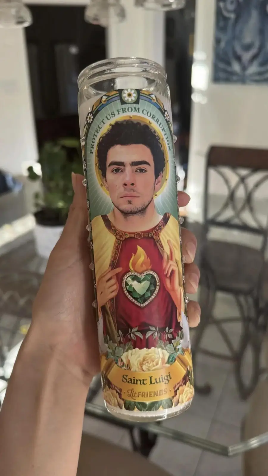 A hand holds a tall, clear glass prayer candle with a printed image of a man resembling Luigi from the Mario video game…