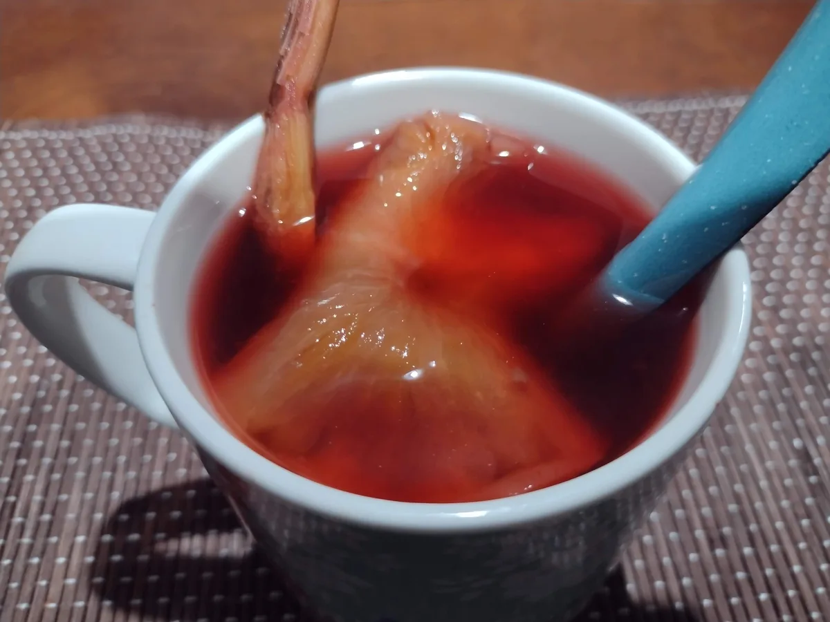 A white ceramic mug sits on a brown woven placemat, filled with a red liquid and containing a large, translucent, gelatinous…