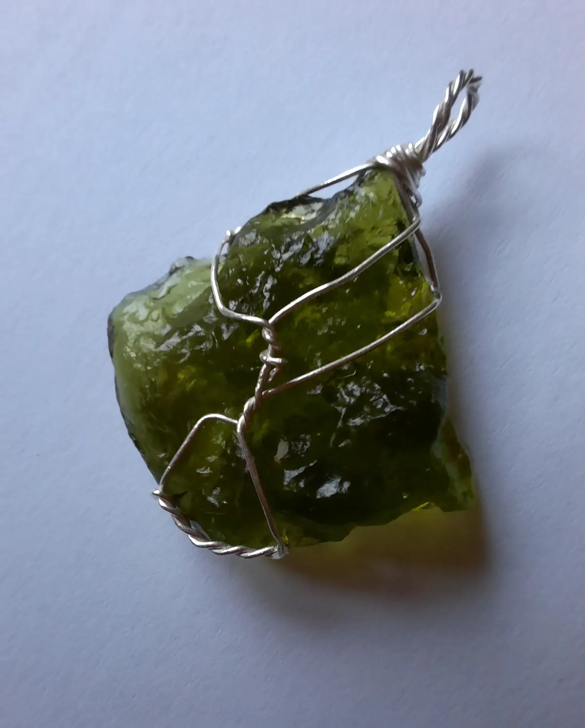 A rough, green, translucent gemstone, likely moldavite, is wrapped in a silver-colored wire frame that forms a loose…