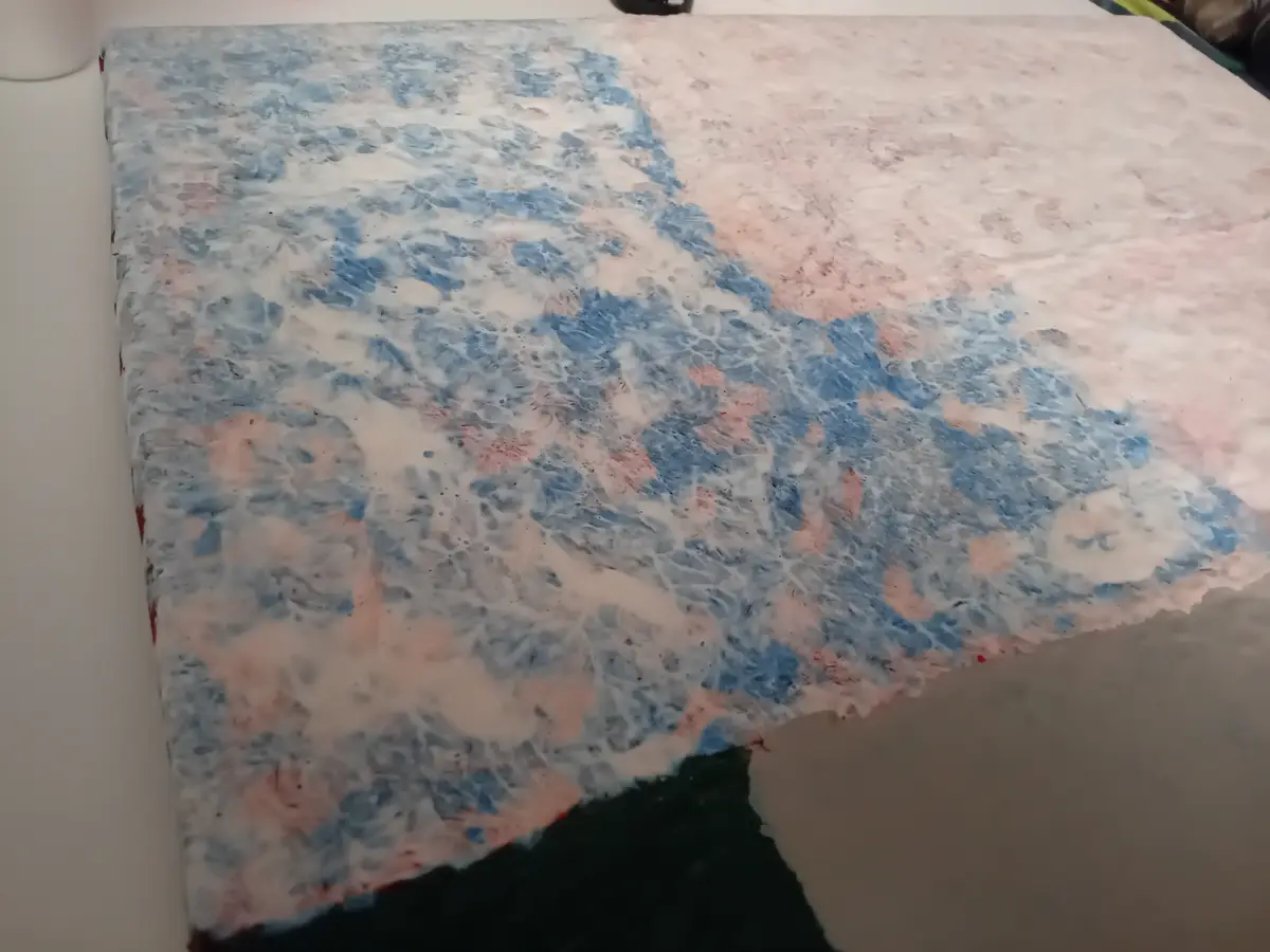 A close-up photo shows a rectangular canvas with abstract, swirling patterns of blue, pink, and white paint, resembling a…
