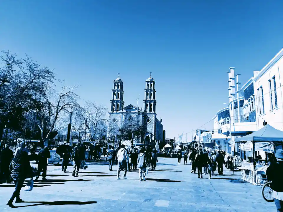 A blue-tinted photo of a busy public square with people walking, featuring a prominent church with two towers in the…