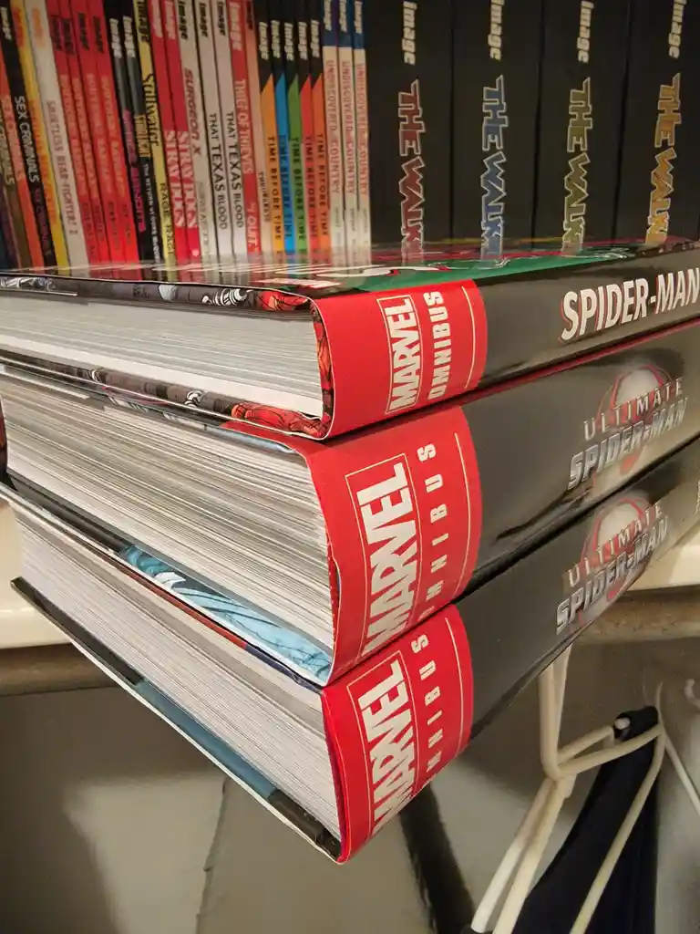 A close-up shot shows three thick, oversized Marvel Omnibus comic books stacked vertically, with 'SPIDER-MAN' visible on the…