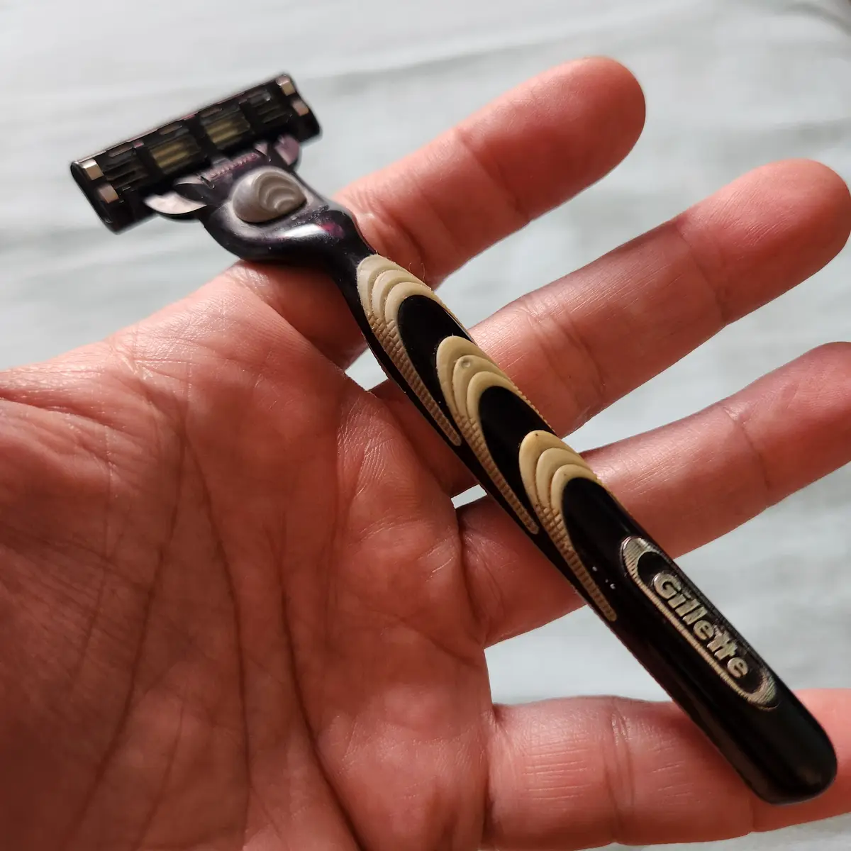 A close-up photo shows a person's hand holding a black and beige Gillette razor, with the brand name visible on the handle…