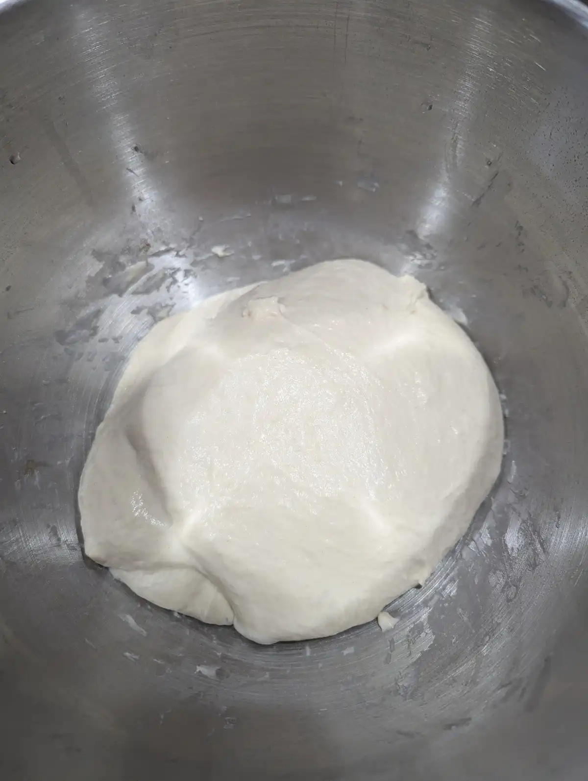 A close-up photo shows a smooth, risen, pale white dough resting in a large, scratched stainless steel mixing bowl. The…