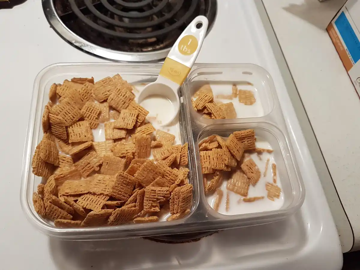 A clear plastic divided food container sits on a white stovetop, holding three compartments filled with cereal and milk. The…