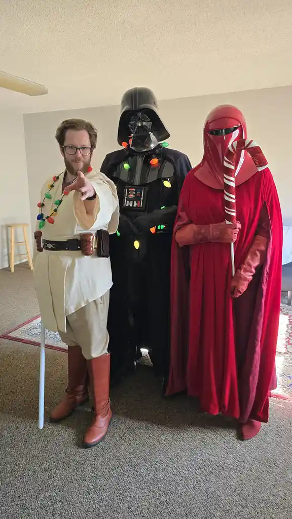 Three people dressed as Star Wars characters pose indoors: a man in a Jedi robe with a lightsaber and Christmas lights…