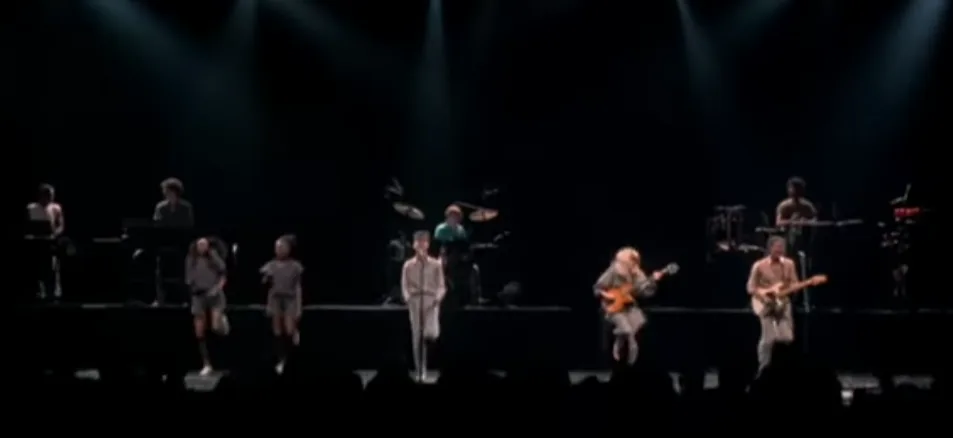 A band performs on a dark stage under spotlights, with the lead singer center stage and musicians around him. Two dancers in…