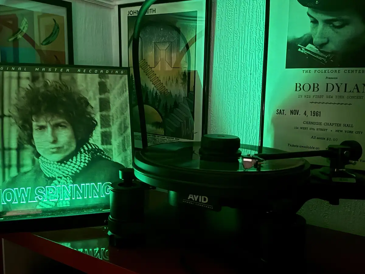 A green-lit room features a turntable with a record on it, surrounded by framed posters including one of Bob Dylan and…