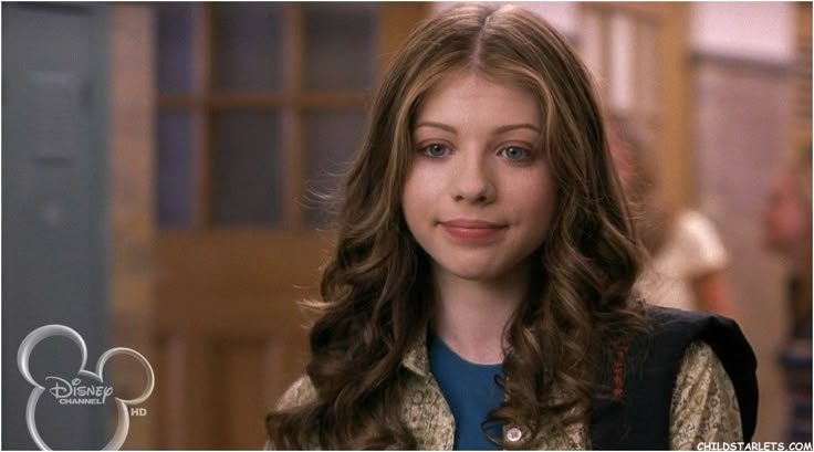 A young woman with long brown wavy hair and a subtle smirk looks directly at the camera. She wears a patterned shirt under a…