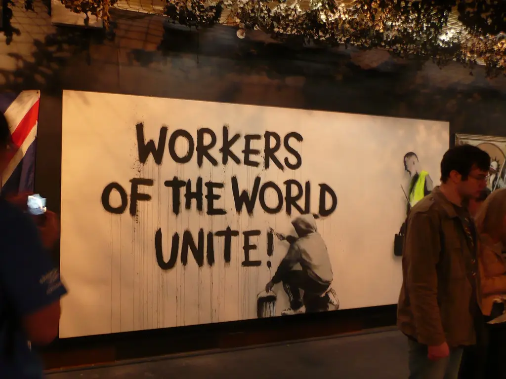 A large white banner displays the phrase 'WORKERS OF THE WORLD UNITE!' in bold black graffiti-style lettering, with a…