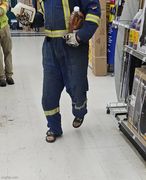 A person in blue work overalls with yellow reflective stripes walks through a hardware store aisle, holding two large…