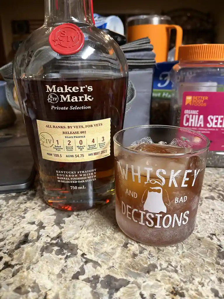 A bottle of Maker's Mark Private Selection bourbon sits next to a glass filled with whiskey and ice. The glass is etched…