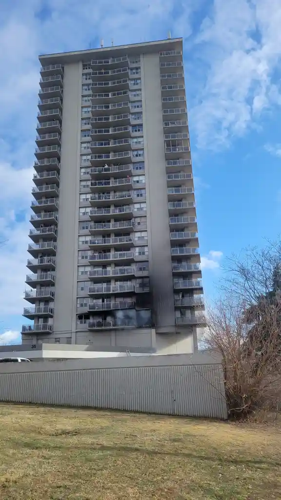 A tall, grey apartment building with numerous balconies under a blue sky with scattered clouds. Smoke is visibly billowing…