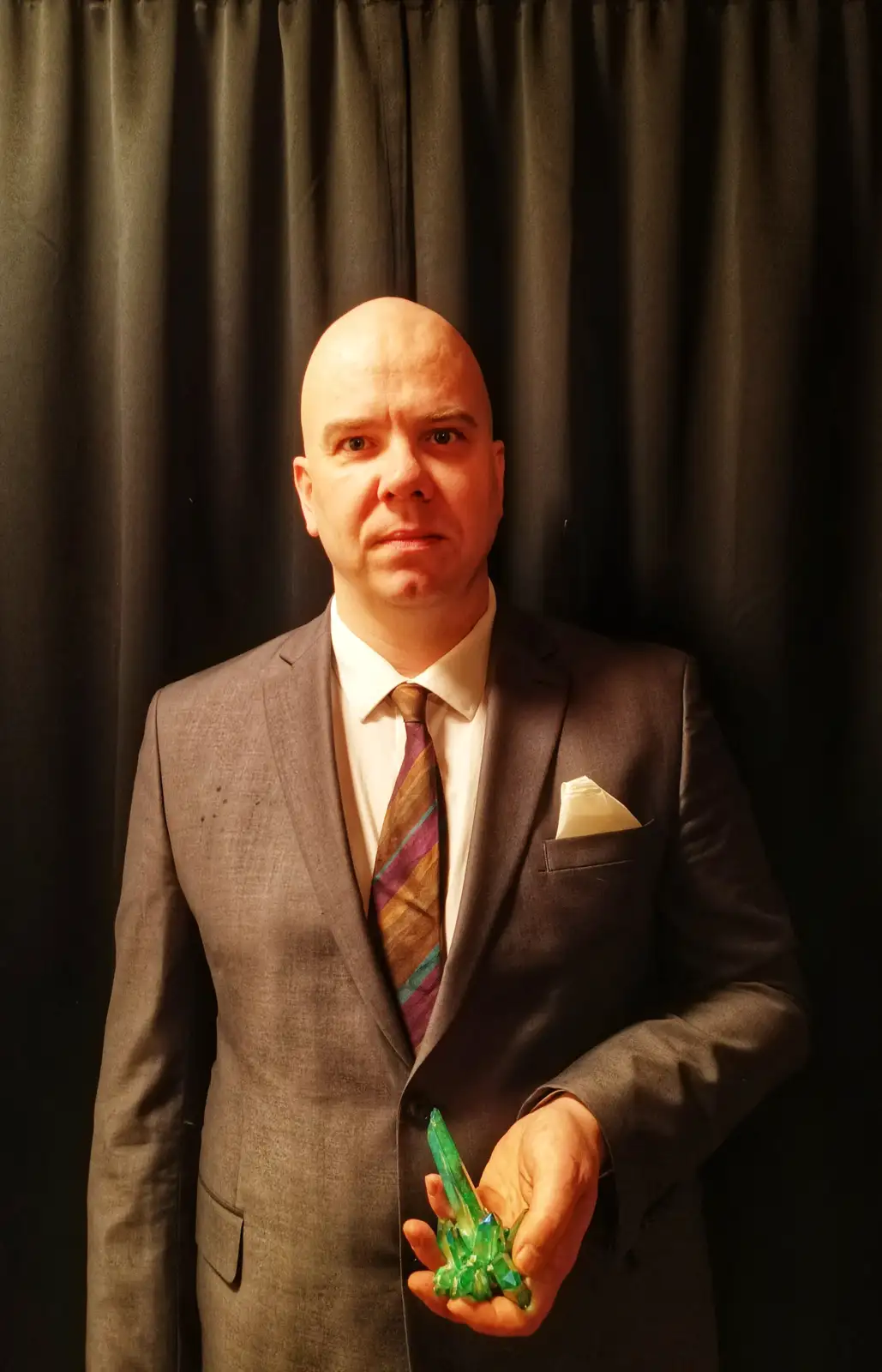 A bald man in a grey suit and striped tie stands against a dark curtain, holding a green crystal cluster in his right hand…