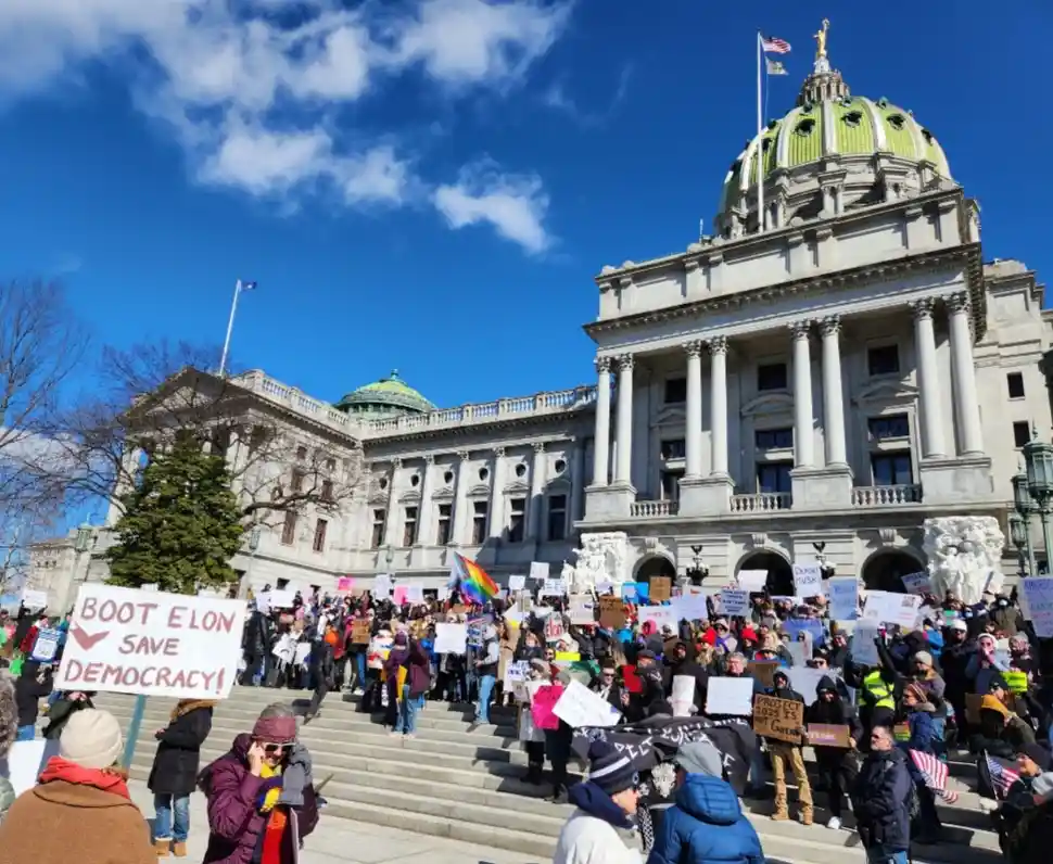 A large crowd of protesters gathers on the steps of a grand neoclassical state capitol building under a bright blue sky with…