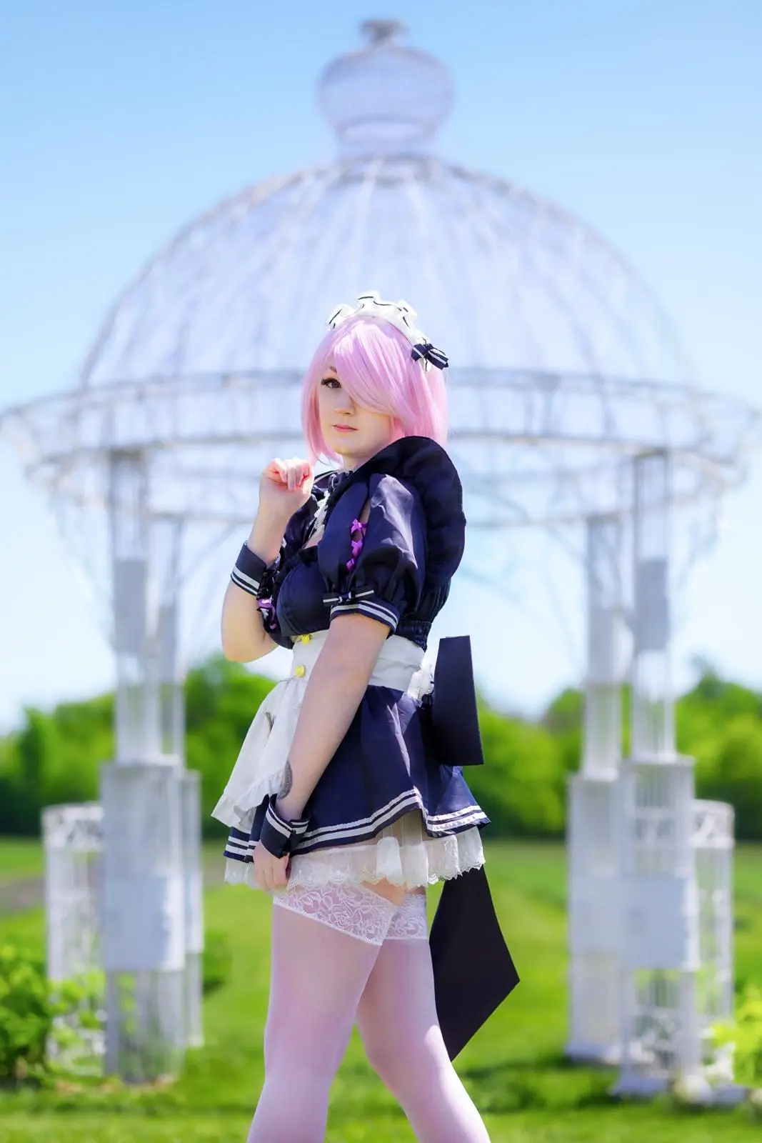 A person with pink hair and a maid outfit poses outdoors in front of a white metal gazebo. The outfit includes a navy blue…