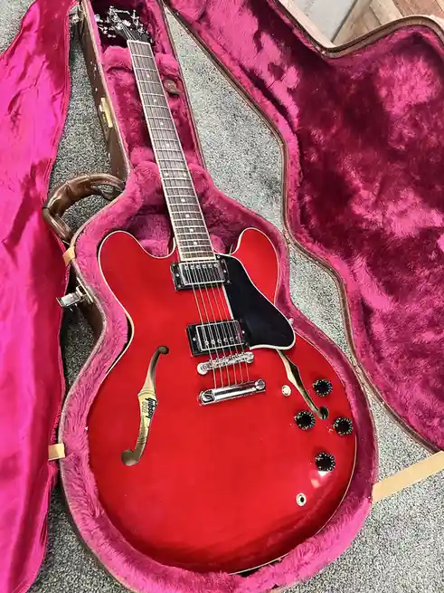 A red Gibson electric guitar with f-holes rests inside an open, pink-furred case on a gray carpet. The overlaid text reads…