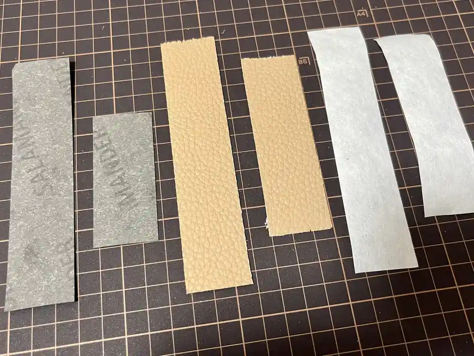 Six rectangular strips of material lie on a grid-lined cutting mat: two gray felt-like strips with faint text, two tan…