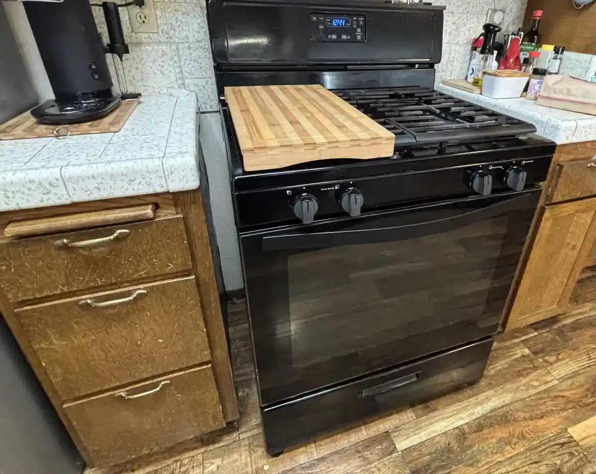 A black gas stove with a wooden cutting board placed directly on its cooktop, surrounded by kitchen cabinets and…