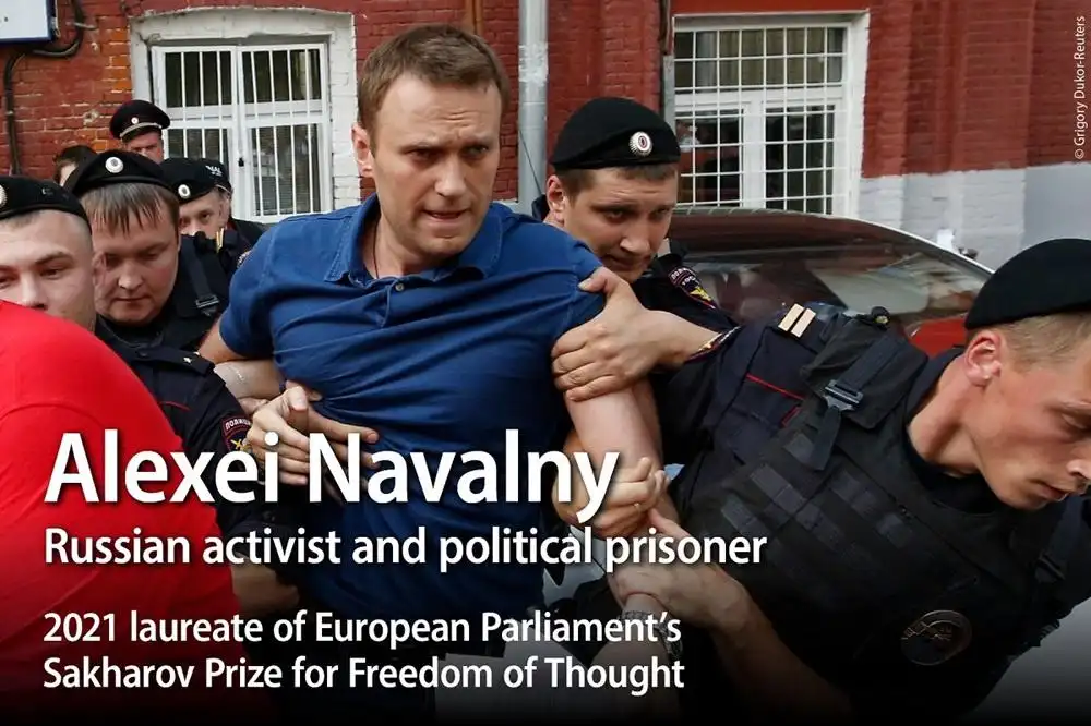 A man in a blue polo shirt is being forcibly restrained by multiple uniformed police officers against a red brick building…