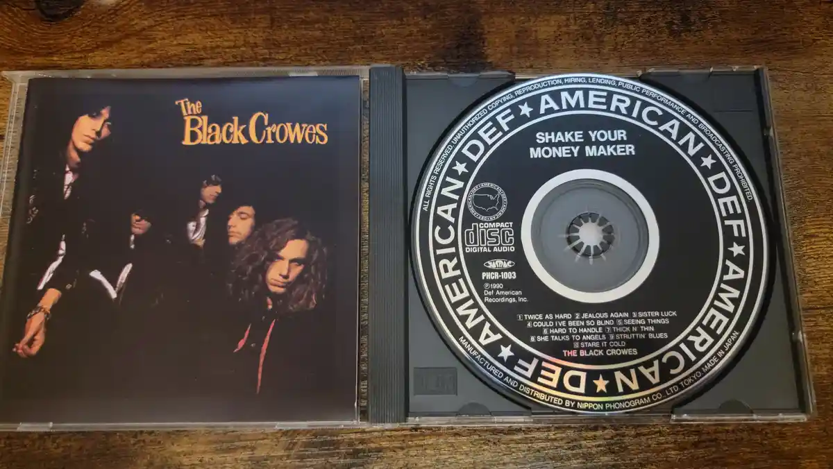 An open CD case displays The Black Crowes' 1990 album 'Shake Your Money Maker'. The left side shows the album cover with the…
