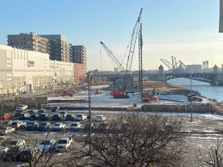 A wide shot of a snowy urban construction site with large cranes, parked cars, and a river with a bridge in the background…