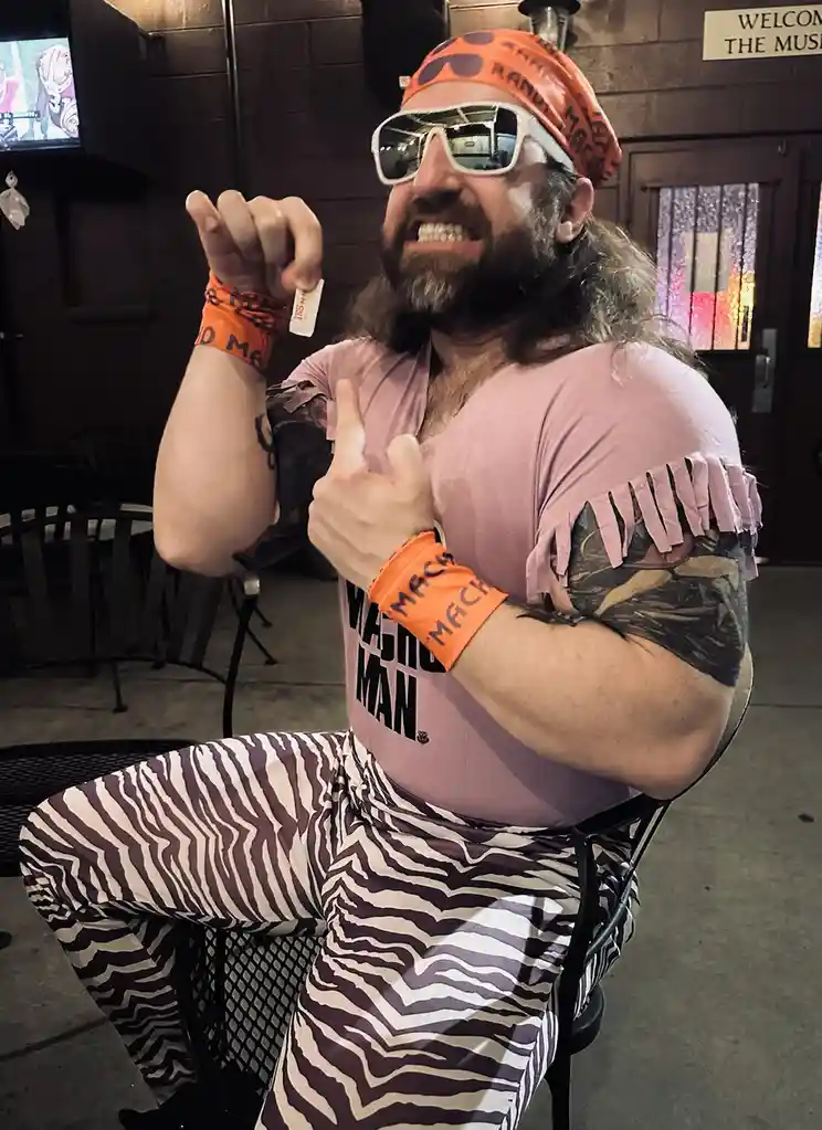 A man dressed in flamboyant wrestling attire, including a pink shirt with 'Macho Man' text, zebra-print pants, orange…